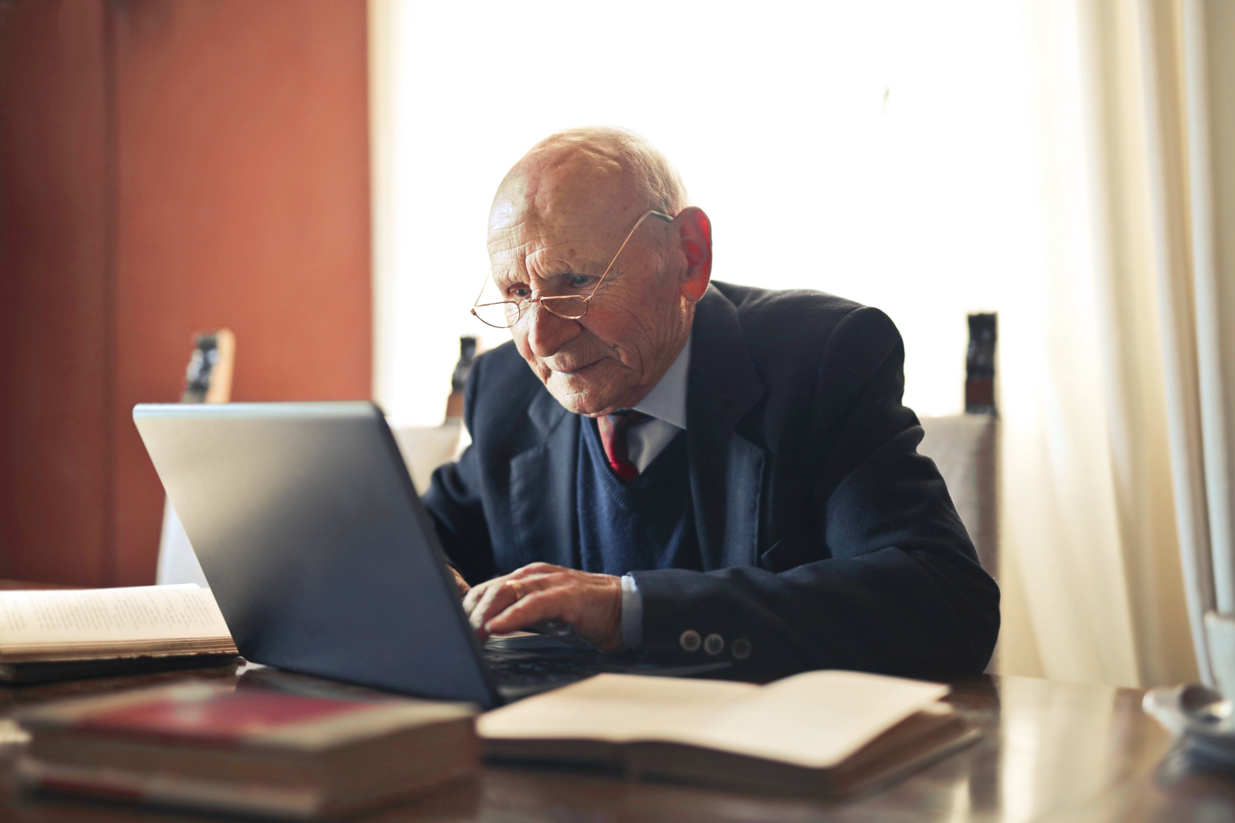 Old man on his home office working on his computer with an open book in front of him