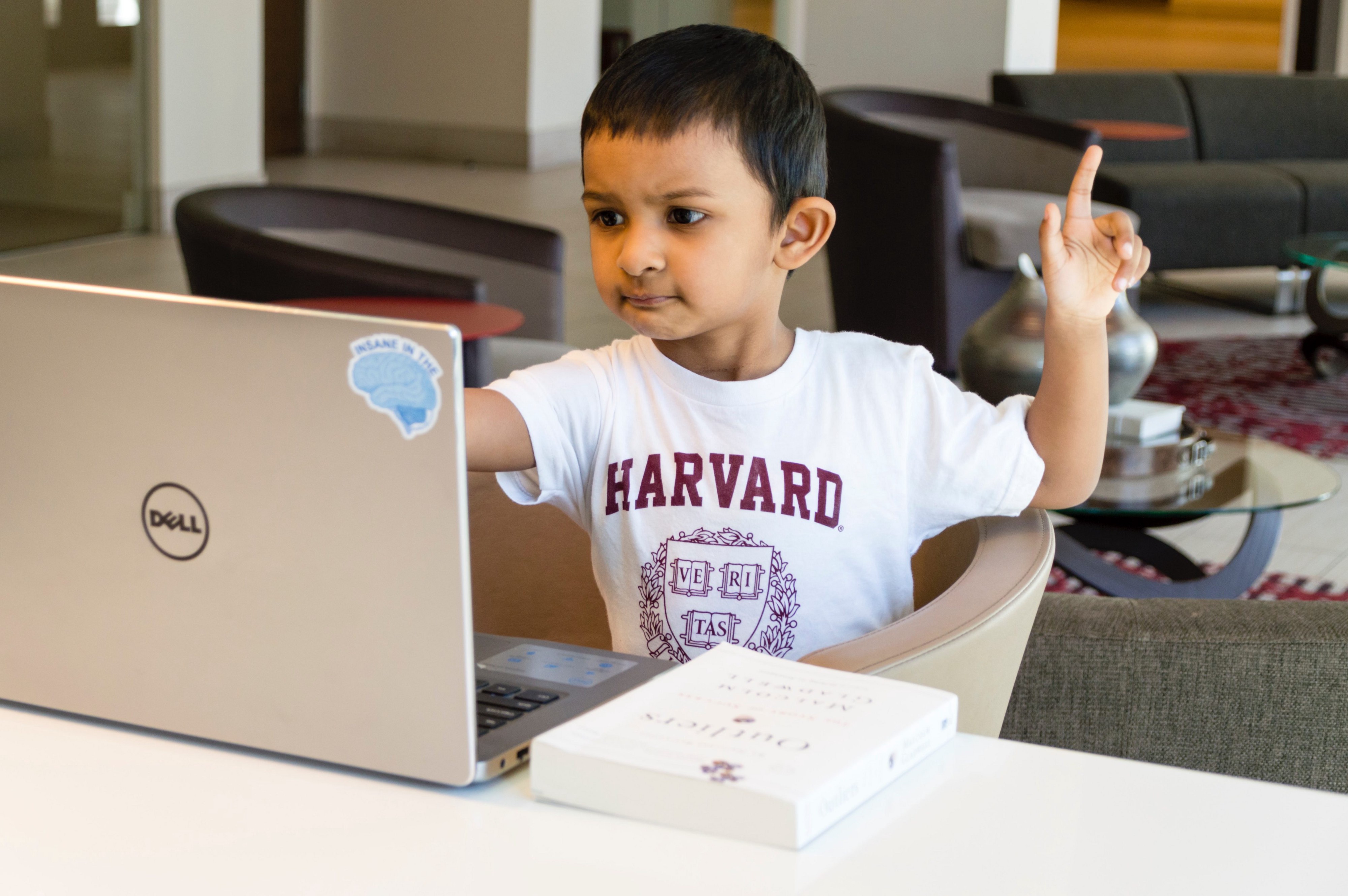 Young kid rising his hand while watching his personal computer with a closed book on the desk.